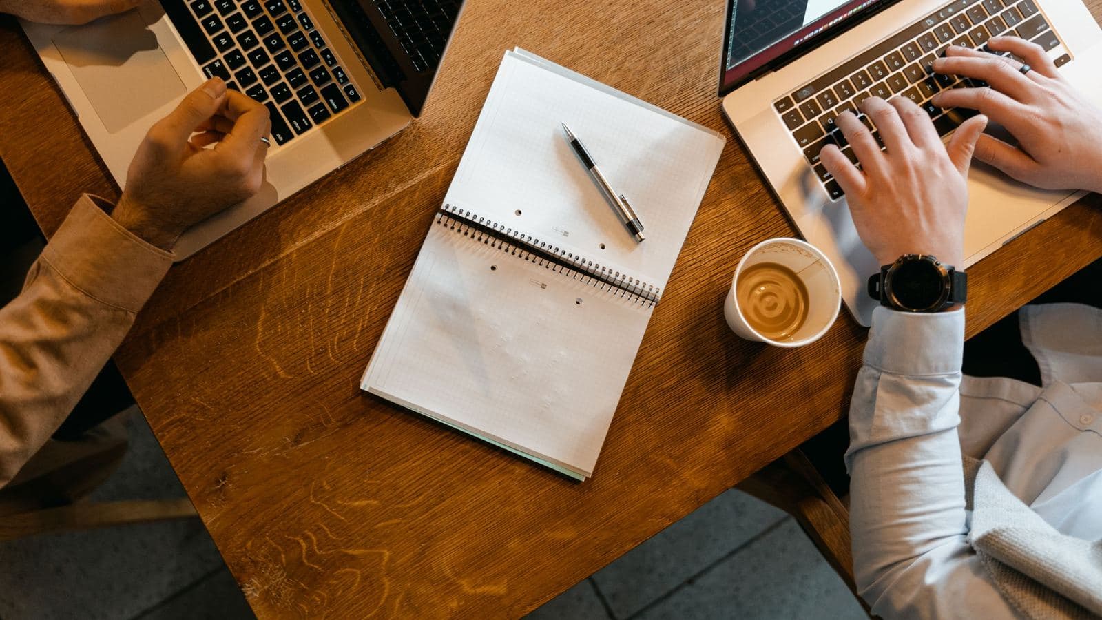 Overhead view of two coworkers typing on laptops at a wooden table, with a notebook and a cup of coffee between them.