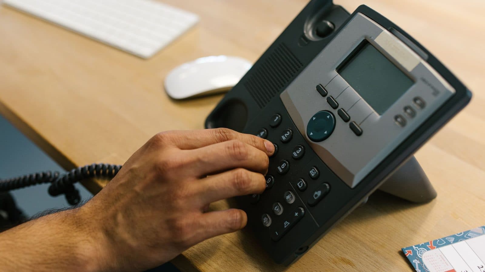 A hand dialing a desk phone on a wooden office desk, next to a keyboard and mouse.