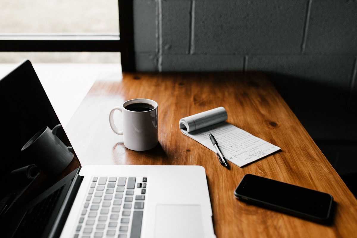 A laptop, a coffee mug, a phone and a notepad with a pen on a wooden desk.